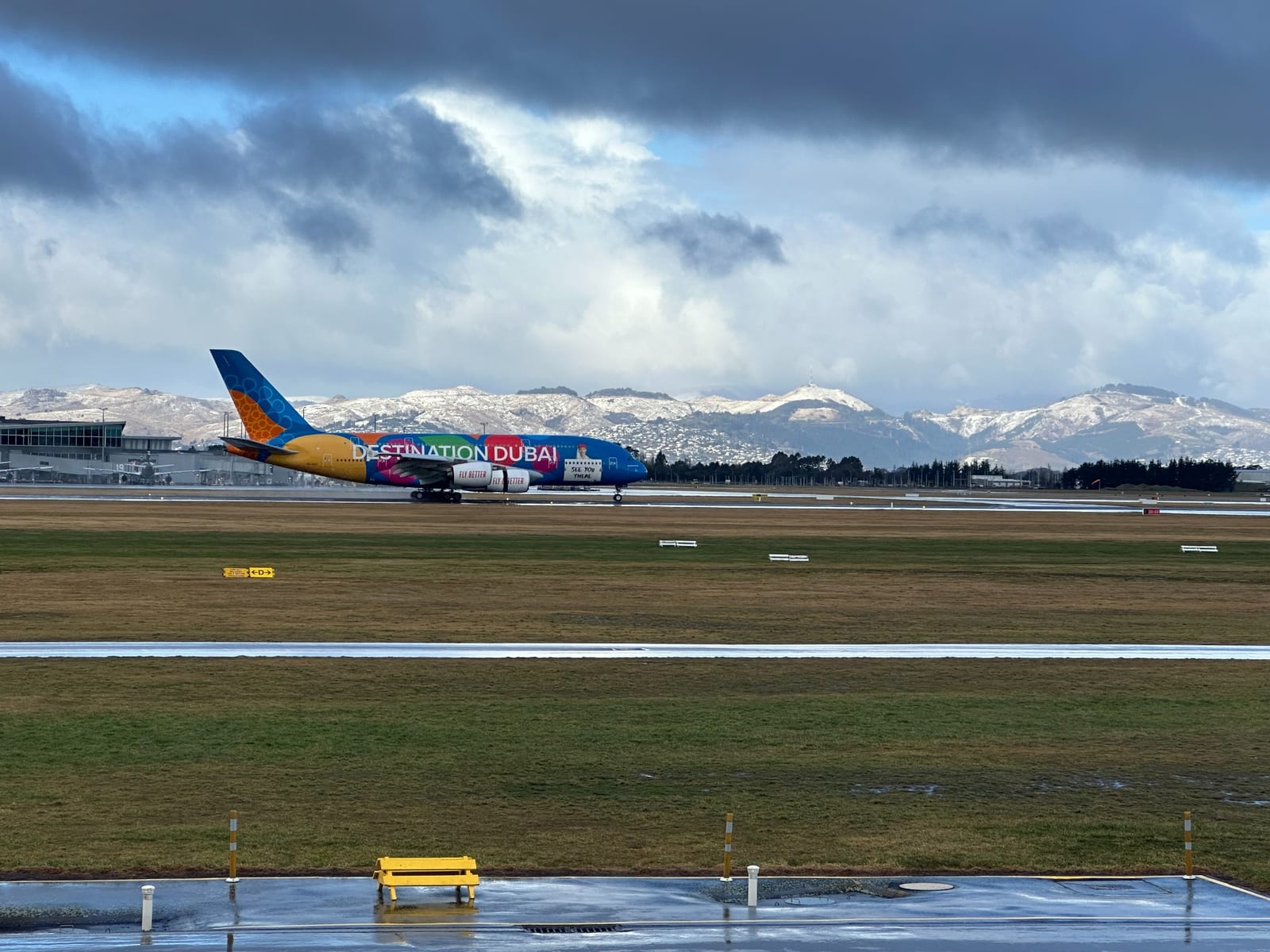 Commercial aircraft on the ground at Christchurch Airport with snow-covered hills and changing weather behind.