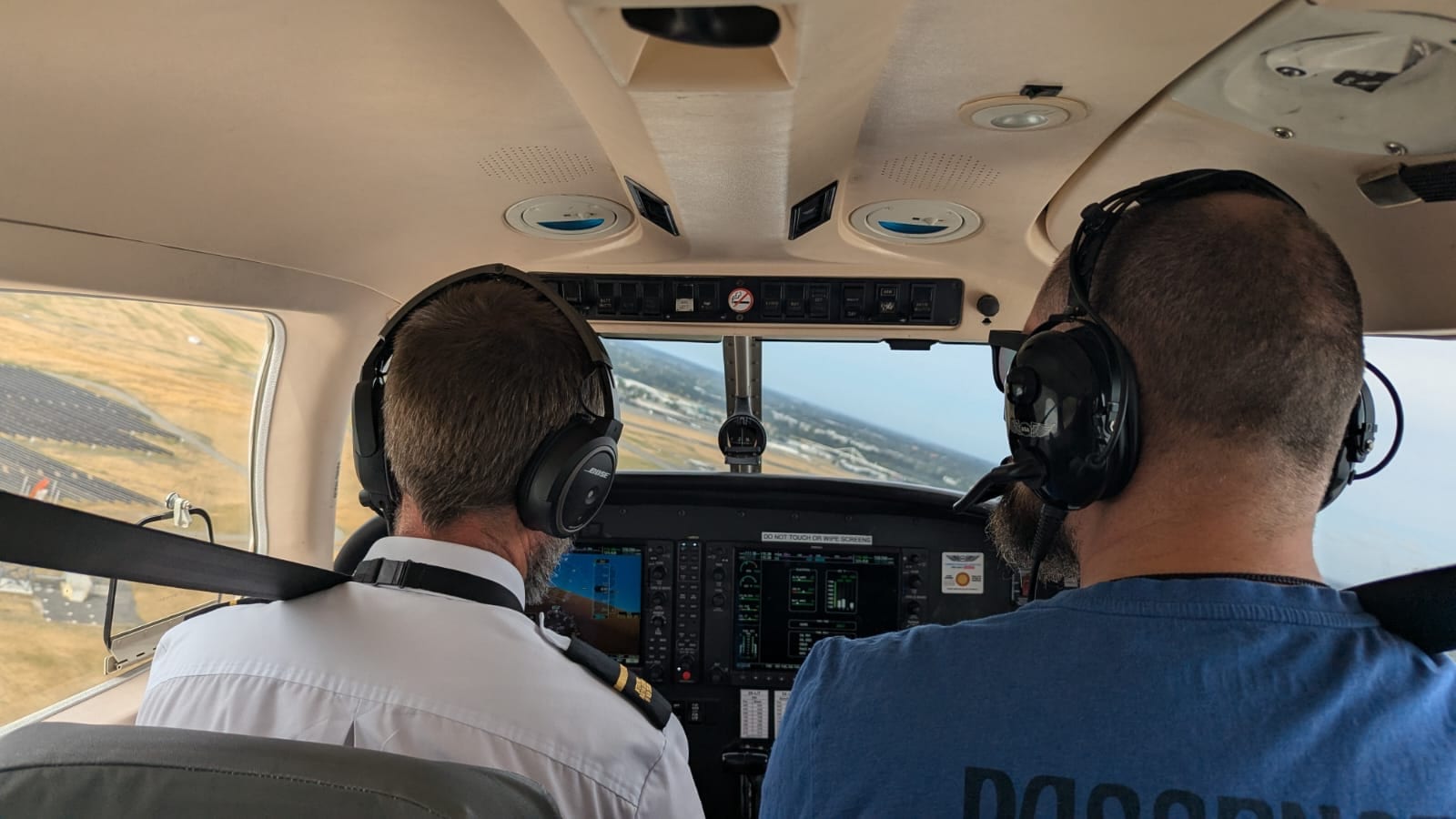 View from the rear seat of a light aircraft cockpit during a training flight with two people wearing headsets.